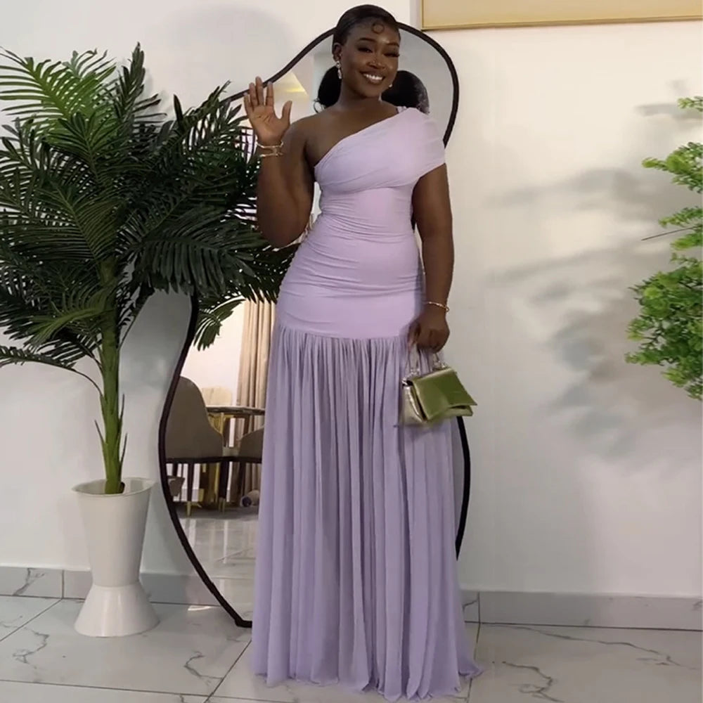 Woman in a lavender one-shoulder dress holding a green handbag indoors.