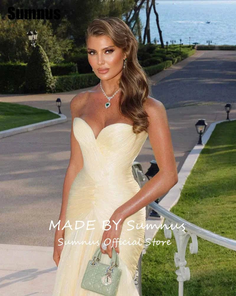 Woman in a strapless yellow dress holding a green handbag, standing outdoors with a scenic background.
