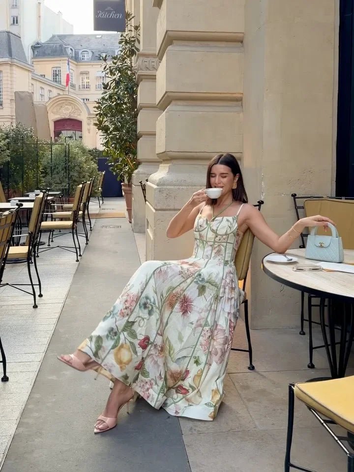 Woman in a floral dress sitting outdoors in an urban setting, drinking from a cup.