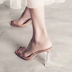 Clear high-heeled shoes worn with a white skirt on a light gray floor.