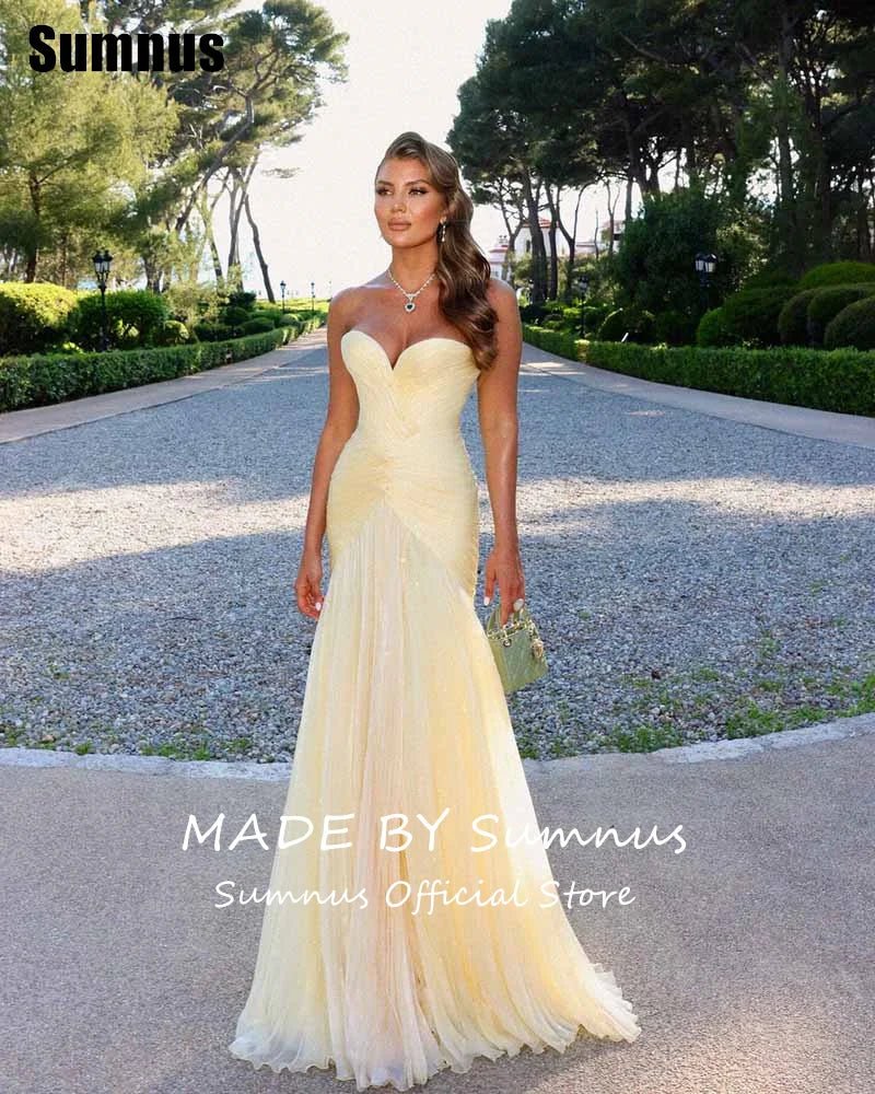 Woman in a yellow evening gown standing on a path with trees and greenery in the background.