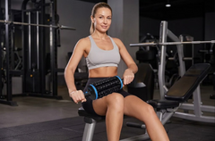 A woman in a gym sits on a bench using a black and blue foam roller on her thigh, wearing a gray sports bra and black shorts.